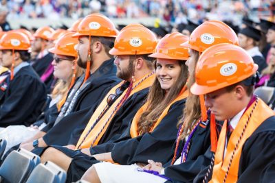 A recent graduate smiles from the commencement ceremony seats while wearing their signature MLSoC hardhats