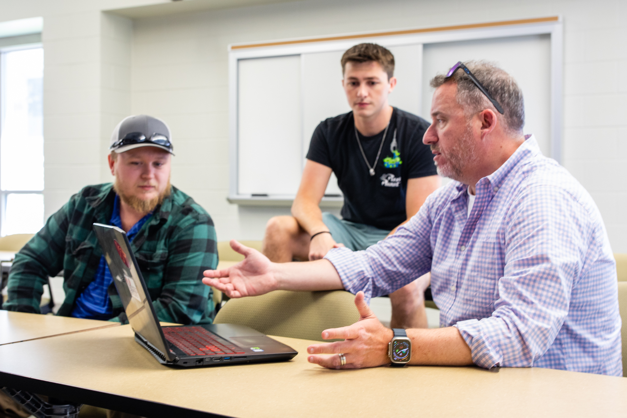 A professor talks with students in a classroom while gesturing to an open laptop on the table.