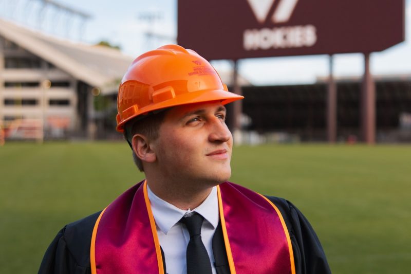 Alex Sherbak, stands on Virginia Tech practice field in an orange hardhat and graduation garb, preparing for career in construction.