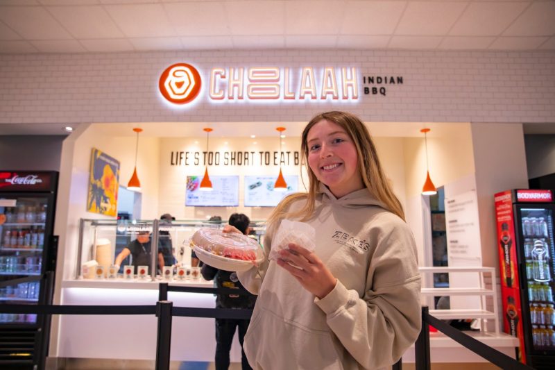 A student holds a to-go container of food and smiles in front of the Choolaah Indian BBQ sign