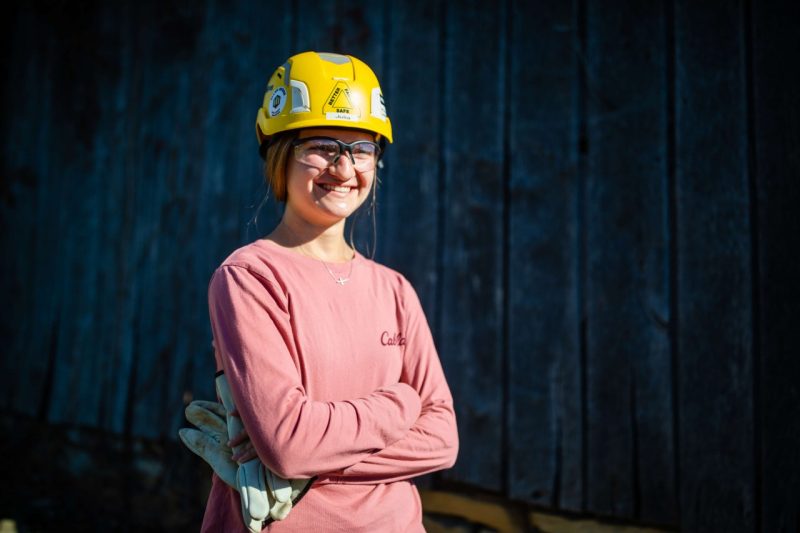 A young woman wearing a hard hat smiling in front of a blue wooden structure.