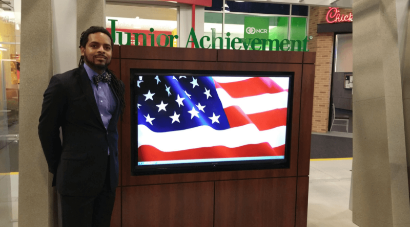 Cosco Jones poses for a photo in front of a screen with an American flag on it and the words "Junior Achievement" in green above.