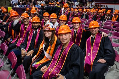 A recent graduate smiles from the commencement ceremony seats while wearing their signature MLSoC hardhats