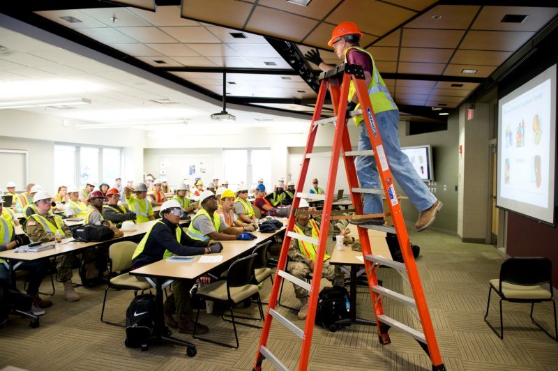 An instructor climbs on a ladder while giving a lecture to a class.