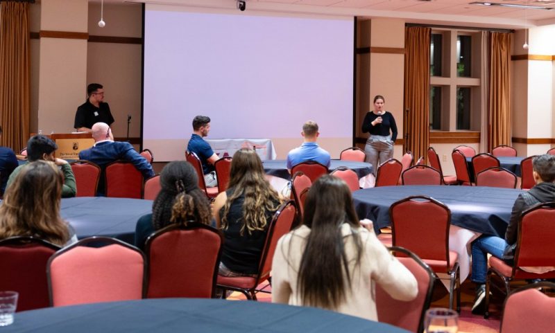 Young Alumni Committee Members Host the Industry Hot Seat Event at the Inn at Virginia Tech with students at circular tables and one woman with a microphone and a man behind a podium.