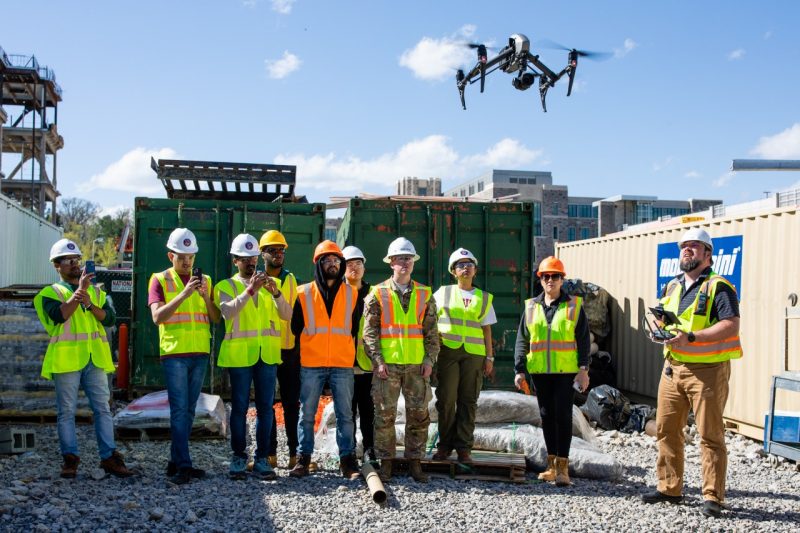 Students on a construction site watch as an instructor flies a drone.