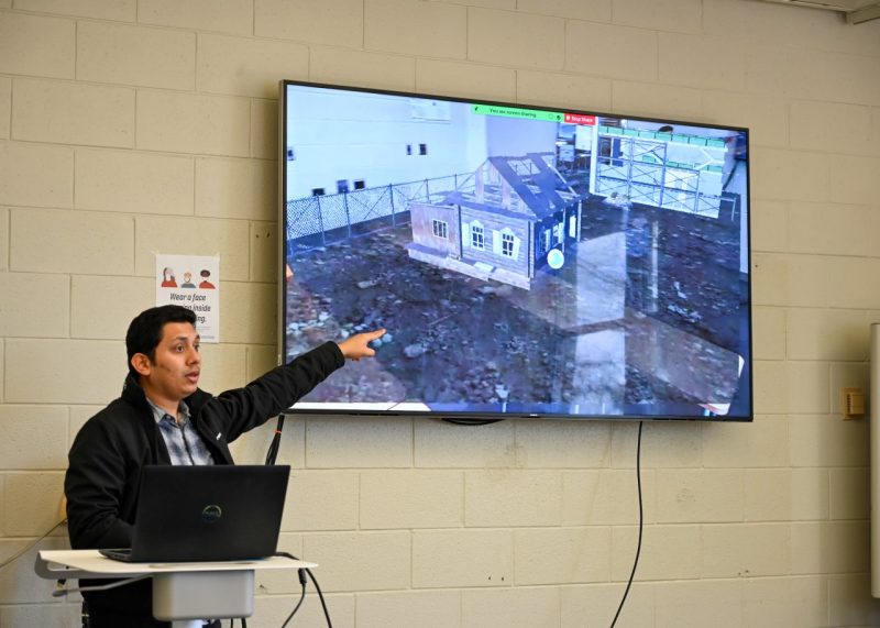 An instructor points to a digital display with a VR view while teaching from behind a laptop on a podium