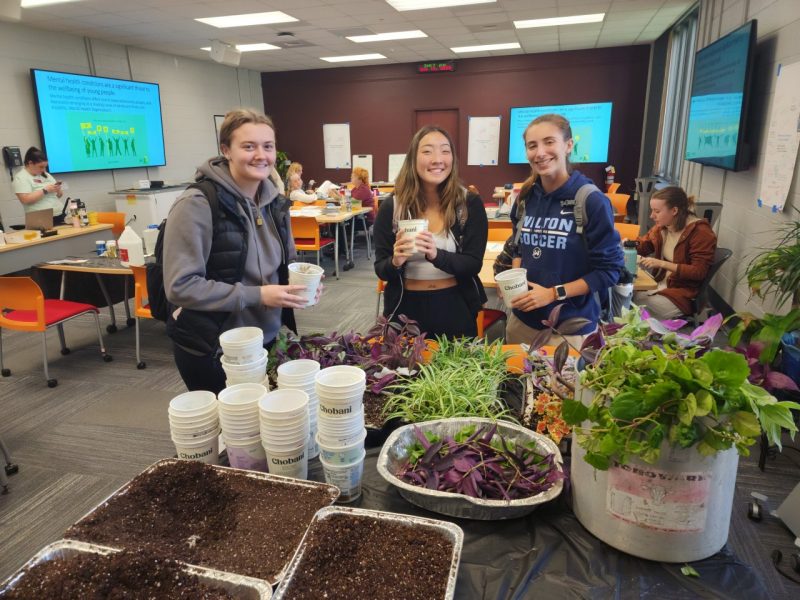 Biobuild students participate in Plantsgiving in a large room with a variety of greenery and empty yogurt containers with three women smiling.
