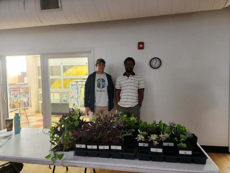 Two students, one with a BioBuild shirt, stand behind a table of free plants to giveaway.