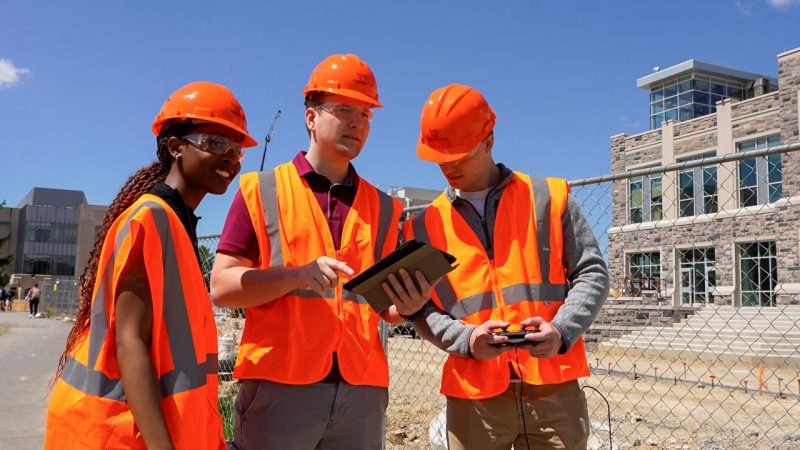 Three students in construction gear look at an iPad while on a construction jobsite.