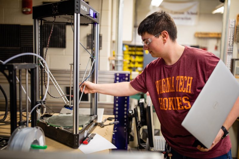 A student wearing a maroon and orange "Virginia Tech Hokies" shirt works with a 3D printing machine.