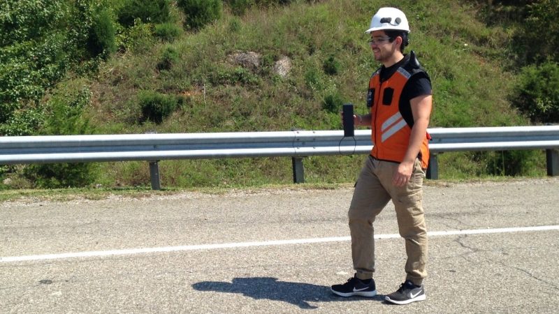 Person wearing a vest walking along a road with a traffic cone