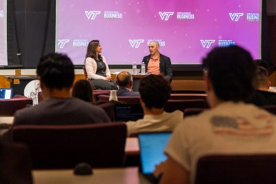 Two people have a discussion in front of a Virginia Tech Pamplin College of Business backdrop in front of a crowd.